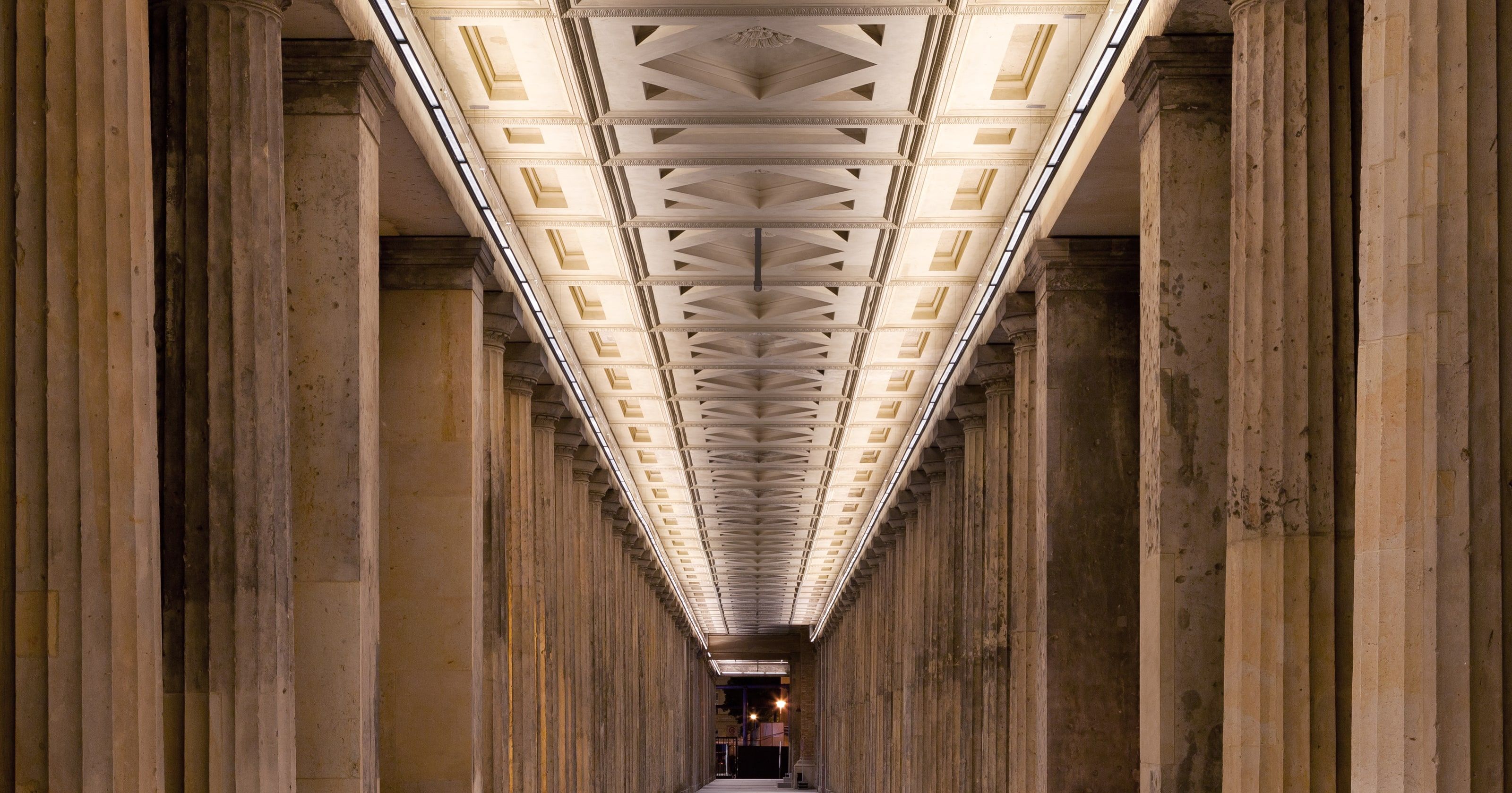 A long stone colonnade with fluted columns and a lit coffered ceiling, receding symmetrically to a distant opening.